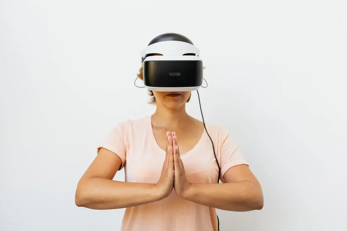 A woman wearing a VR headset meditates with her hands together against a white background.