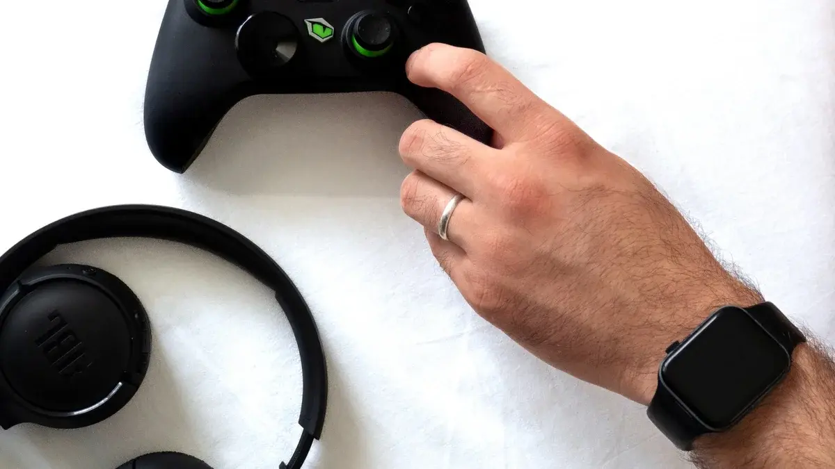 A hand holding a gaming controller next to a headset and smartwatch.