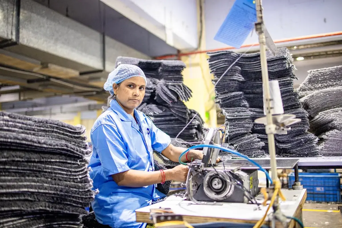 A woman works at an industrial sewing machine in a textile factory surrounded by stacks of fabric.