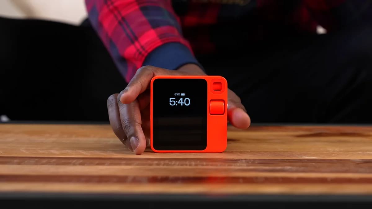 A hand holding an orange Rabbit r1 AI assistant device on a table.