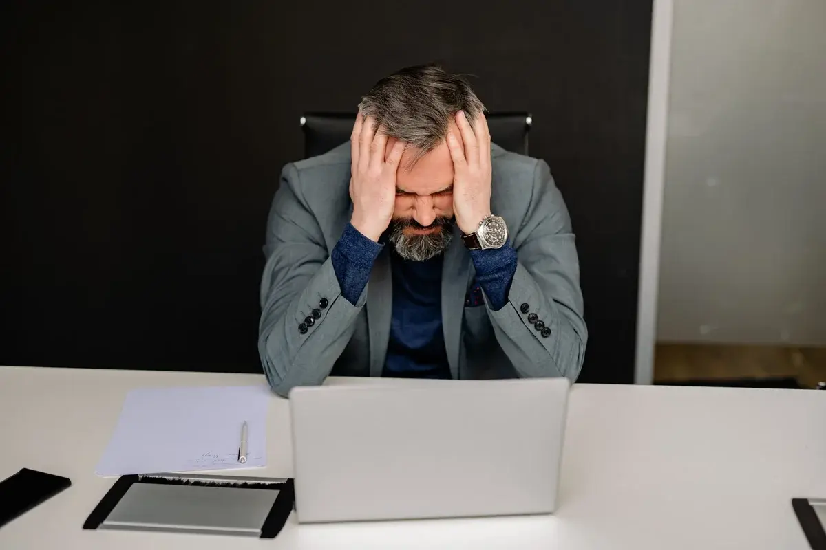 A frustrated businessman holding his head while sitting at a laptop in an office.
