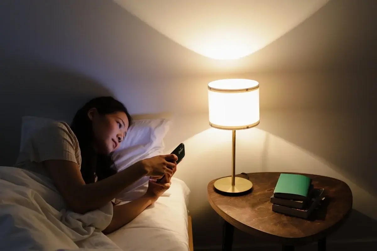 Woman lying in bed using a smartphone beside a lit lamp.