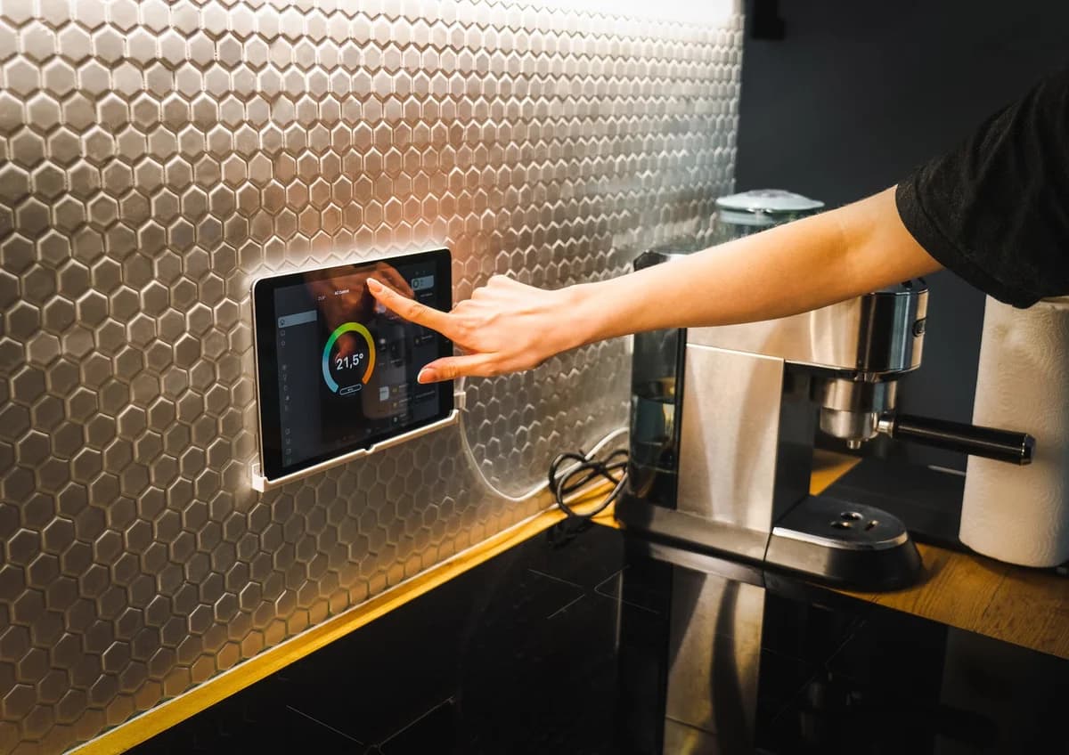 A hand taps a smart home tablet in a modern kitchen.
