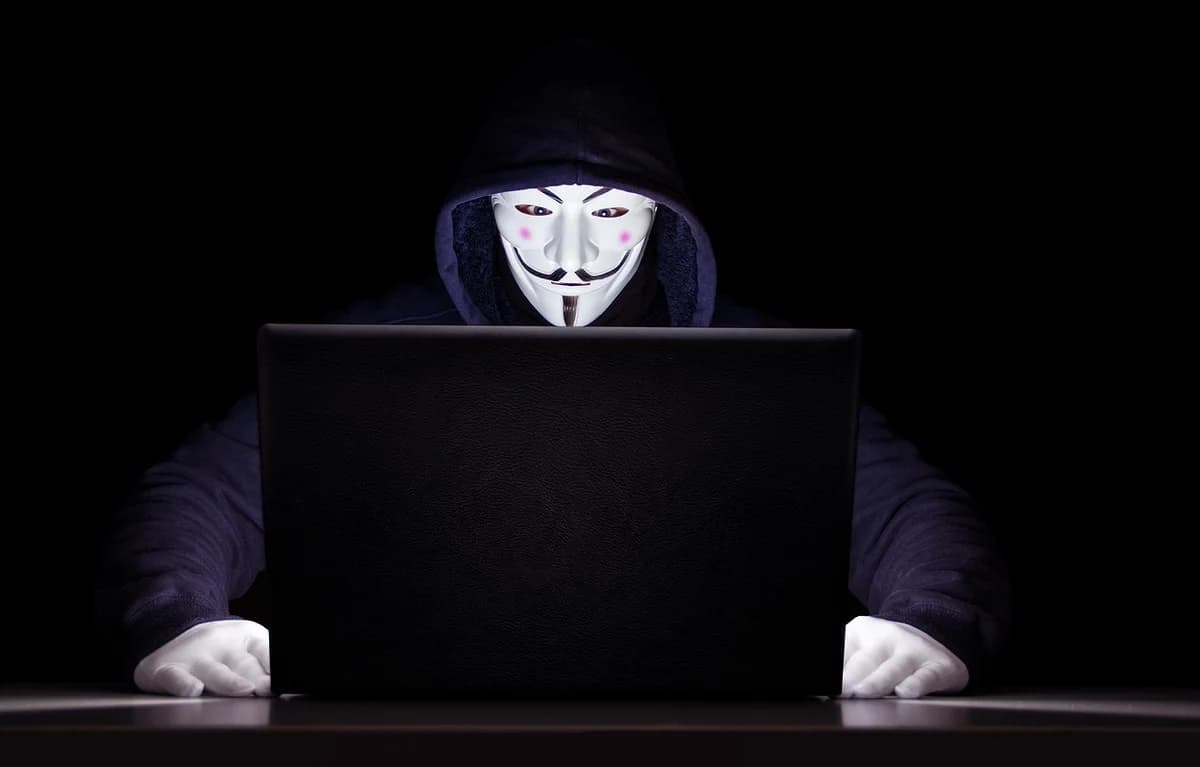 A masked hooded figure using a laptop in the dark.