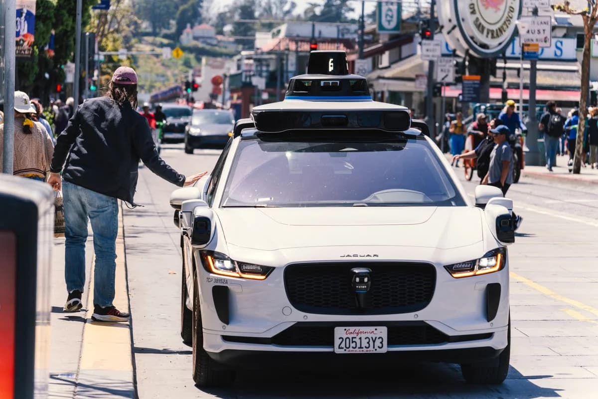 A white self-driving Jaguar car with sensors on top drives through a busy city street near pedestrians.
