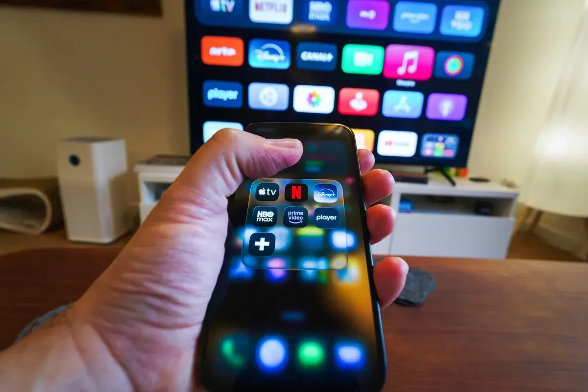 Hand holding smartphone showing streaming app icons with TV screen in background