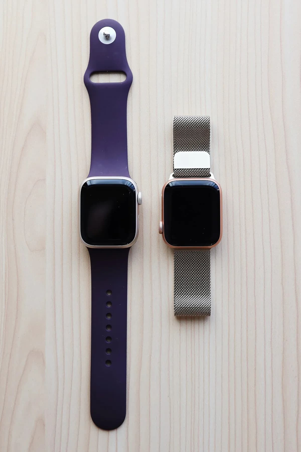 Two Apple Watches on a wooden surface, one with a purple band and one with a mesh band.