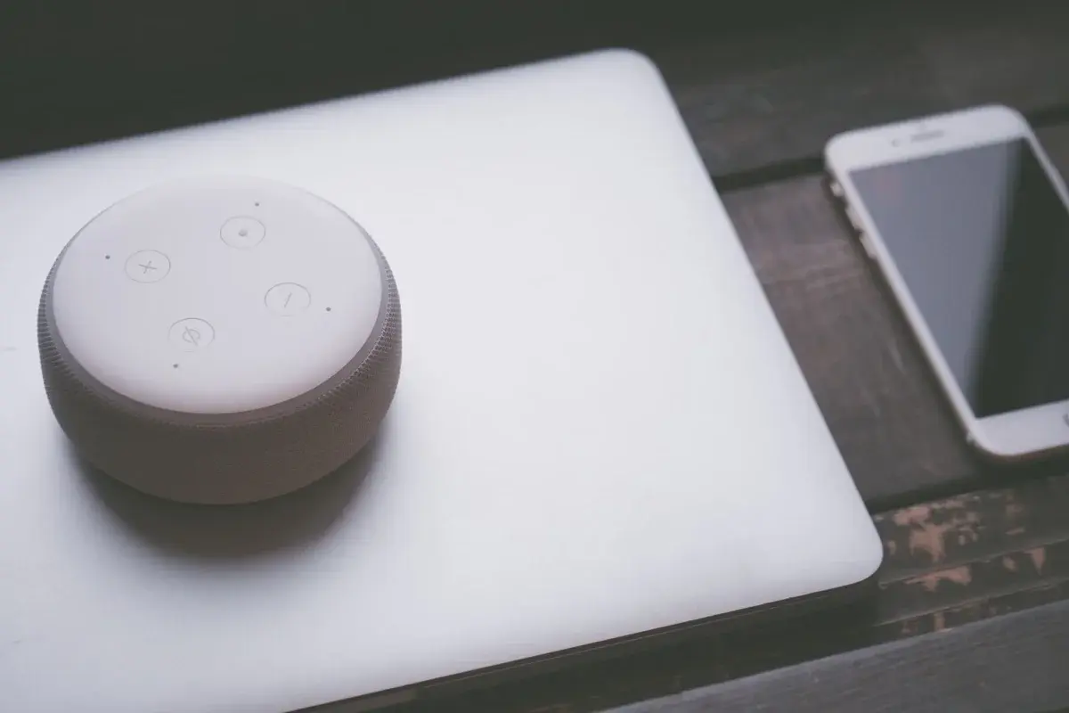 A smart speaker, laptop, and smartphone on a wooden table.