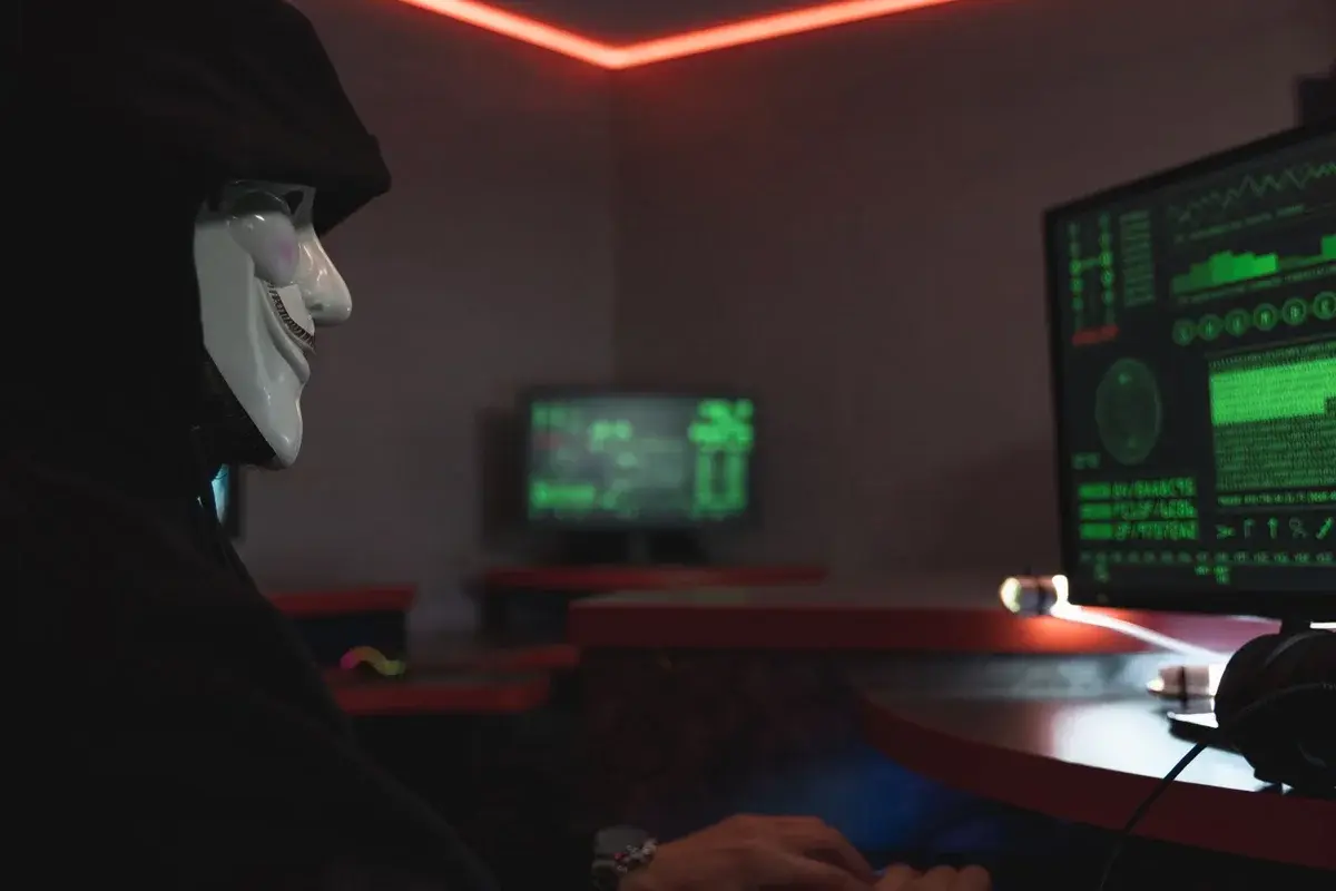 Masked person at a computer in a dark room with glowing screens