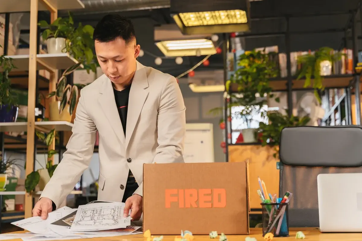 An Asian man in a beige jacket puts papers into a cardboard box labeled "FIRED" in an office.