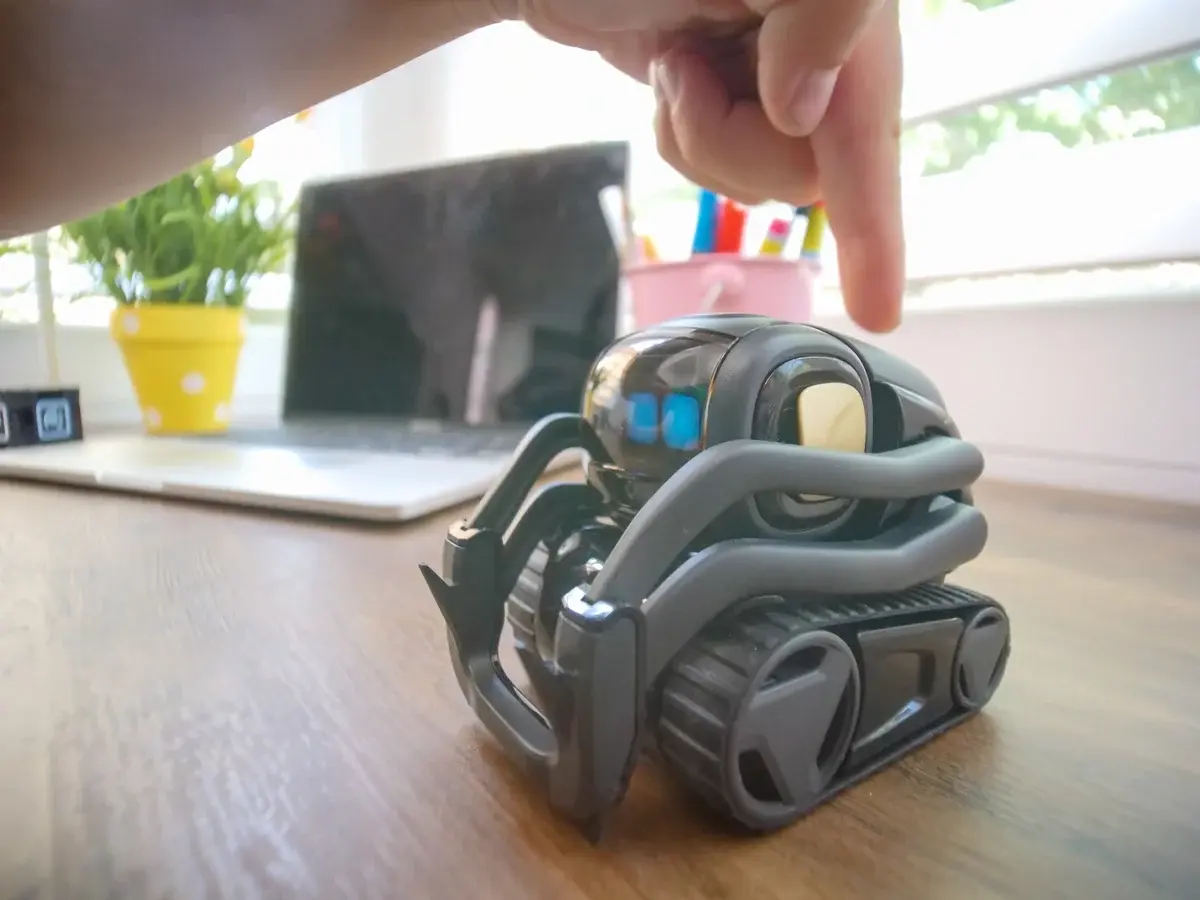 A small interactive robot toy on a desk with a hand pointing at it.